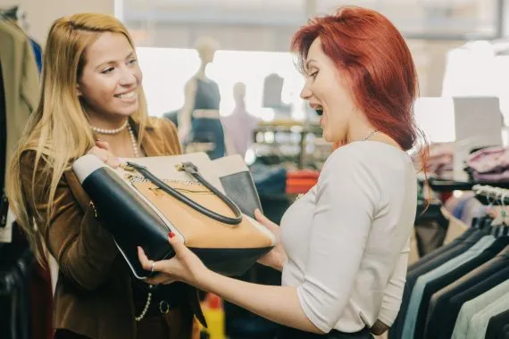 Vendedora de uma loja de roupas sorri enquanto mostra bolsa a uma cliente em uma abordagem de vendas. A cliente pega o acessório em uma reação de surpresa.