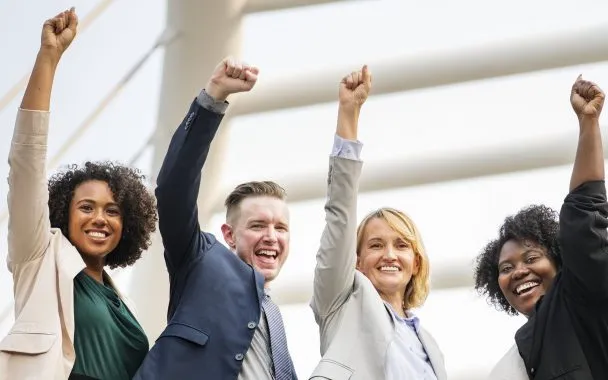Quatro pessoas estão lado a lado em frente a um prédio. Da esquerda para a direita, temos uma mulher, um homem e duas mulheres. Todos estão usando terno, estão sorrindo e têm o punho levantado para o alto em sinal de vitória. A imagem ilustra o tema de como motivar sua equipe.