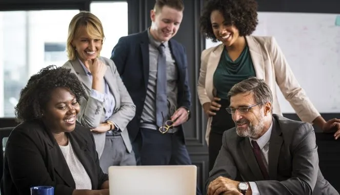 Cinco pessoas estão reunidas em frente a um computador. Três pessoas estão em pé, sendo um homem e duas mulheres, e duas estão sentadas, sendo uma mulher e um homem. A mulher sentada mostra algo na tela do computador para o homem sentado, que parece ser o líder da equipe. Todos sorriem e usam terno. A imagem ilustra o tema de como motivar sua equipe.