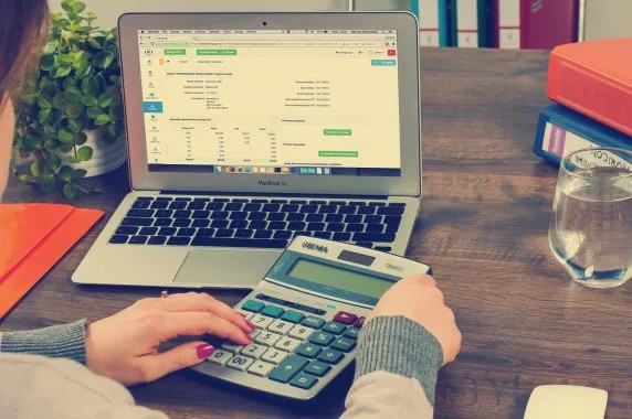 Mulher sentada a uma bancada utilizando calculadora e notebook com ferramenta de gestão, pensando em como organizar as finanças.