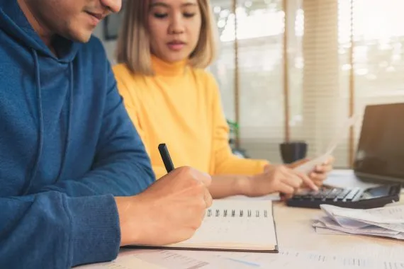 Casal sentado a uma bancada discutindo como organizar as finanças. O homem escreve em um caderno e a mulher separa notas fiscais enquanto usa uma calculadora.