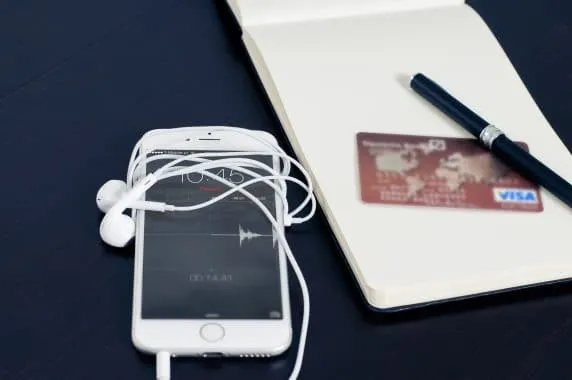 Um celular com fone enrolado está em cima de uma mesa e ao lado de um bloco de notas. O bloco tem um cartão de crédito e uma caneta sobre ele. A aproximação dos objetos faz referência ao comportamento do consumidor imediatista e multicanal.