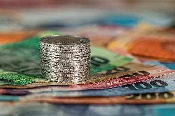 Em uma mesa estão espalhadas várias notas de dinheiro de valores diferentes. Cada valor tem uma cor. Em cima das notas tem uma pilha de moedas. A imagem representa o departamento comercial de uma empresa.