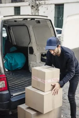 Homem de calça, casaco e boné retira caixas de papelão de furgão com porta aberta. A cena simboliza a importância de entregas regulares para uma boa gestão de fornecedores.