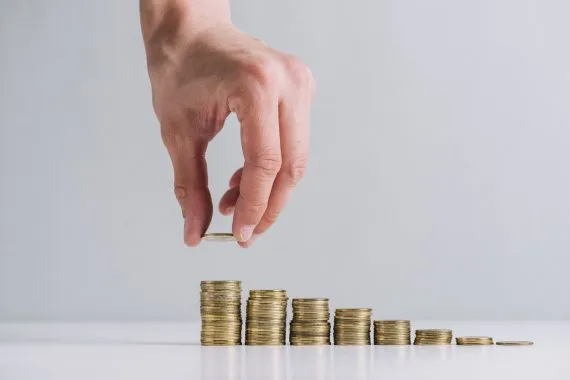 Uma mão masculina coloca moeda na maior de sete pilhas crescentes de dinheiro simbolizando o controle de caixa na gestão financeira.