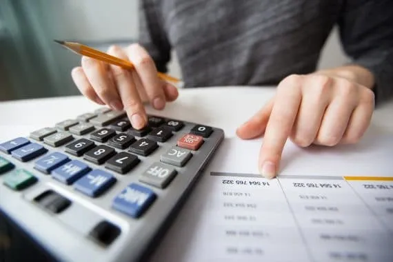 Uma mulher sentada a uma mesa faz a gestão financeira de uma empresa usando calculadora e apontando para uma folha com números