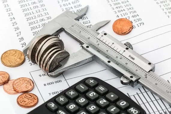Um paquímetro segura uma série de moedas enquanto está apoiado sobre documentos com números. A imagem simboliza uma precificação segura feita por mark up.