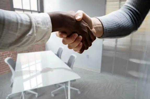 Duas mãos se cumprimentam em primeiro plano como se fechassem uma contratação após fase de recrutamento e seleção. O ambiente é o de um escritório, com uma mesa, três careias e uma janela ao fundo.